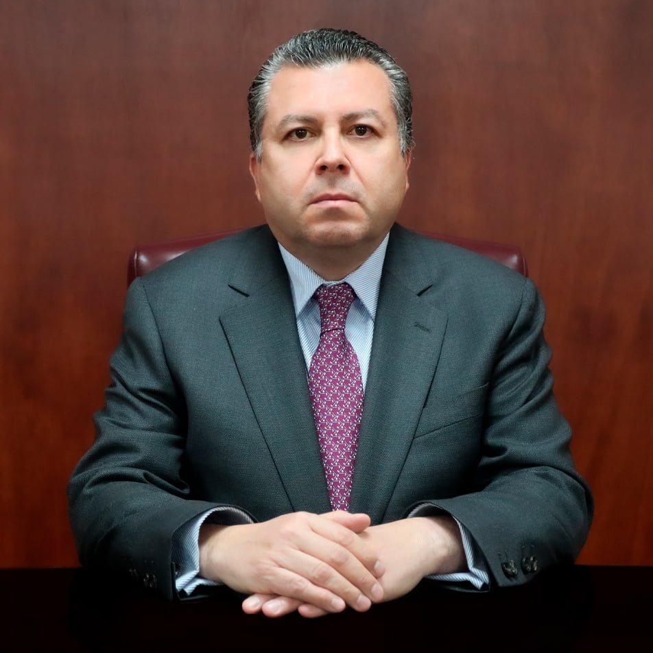 Professional headshot of a middle-aged man in a dark suit and purple tie, seated against a burgundy background with hands folded