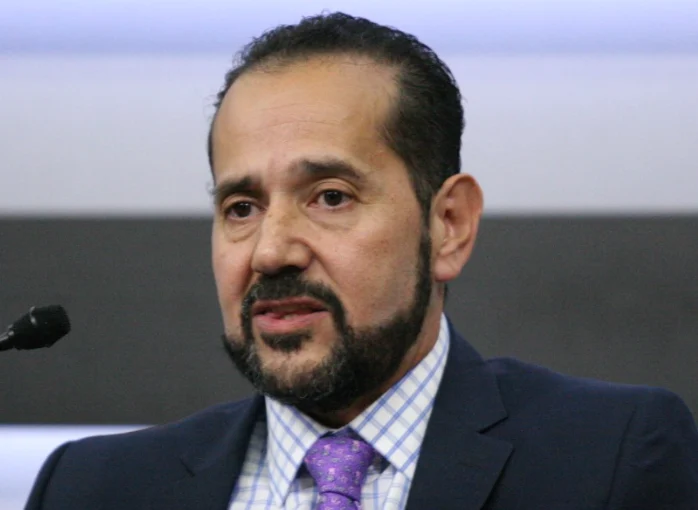 Man with dark hair and beard wearing navy suit and purple tie speaking at microphone