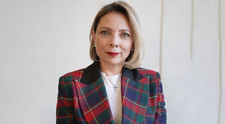 Professional headshot of a woman wearing a red and navy plaid blazer over a white shirt against a neutral background